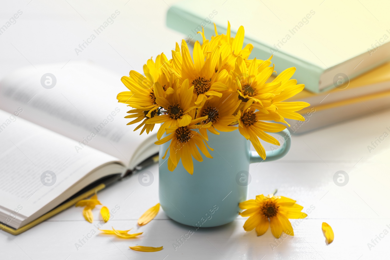 Photo of Beautiful bright yellow flowers in light blue cup near books on white table