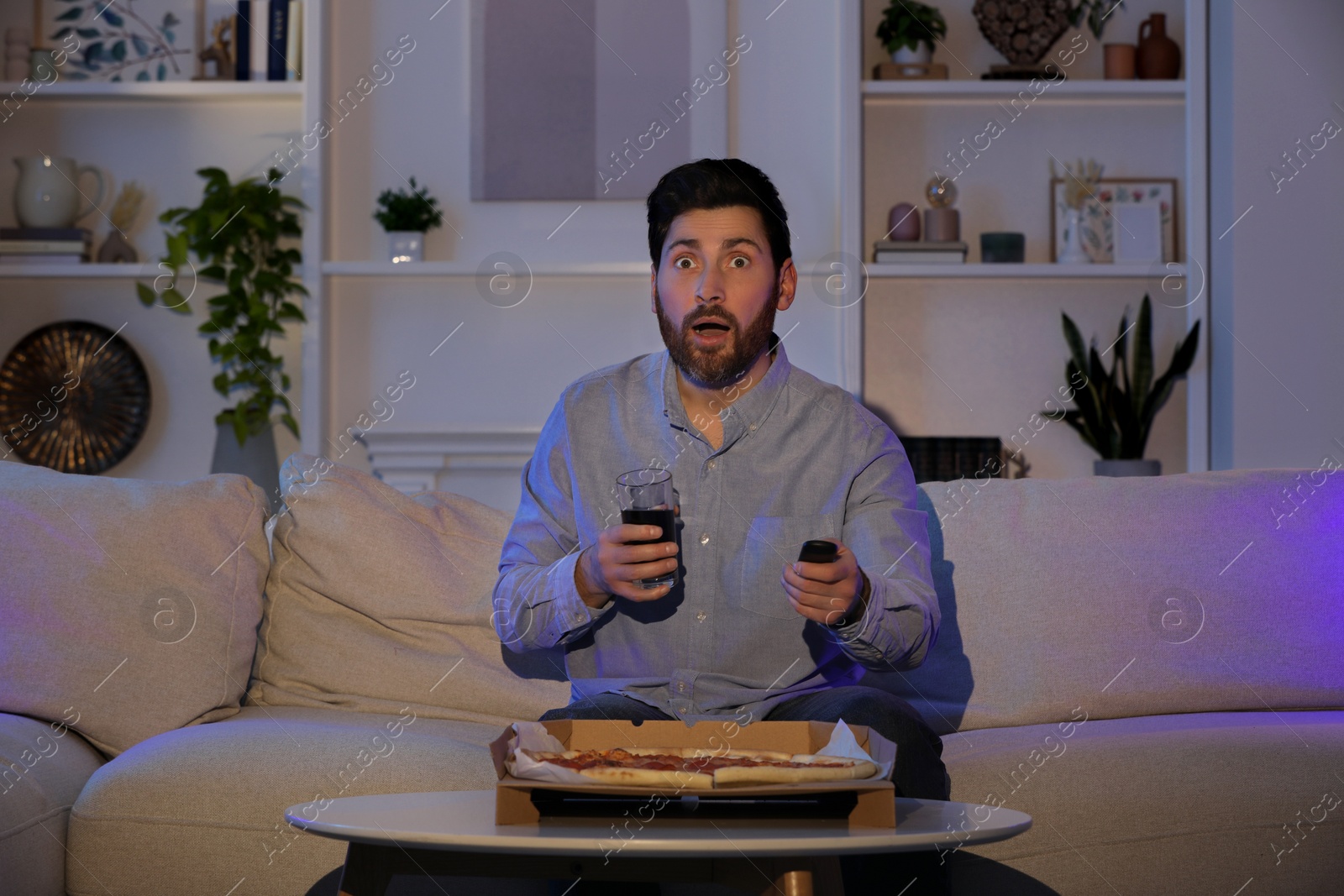 Photo of Frightened man watching TV with pizza on sofa at home