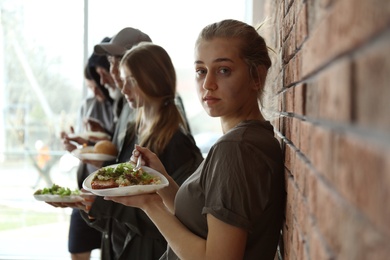 Photo of Young poor woman and other people with food at brick wall indoors