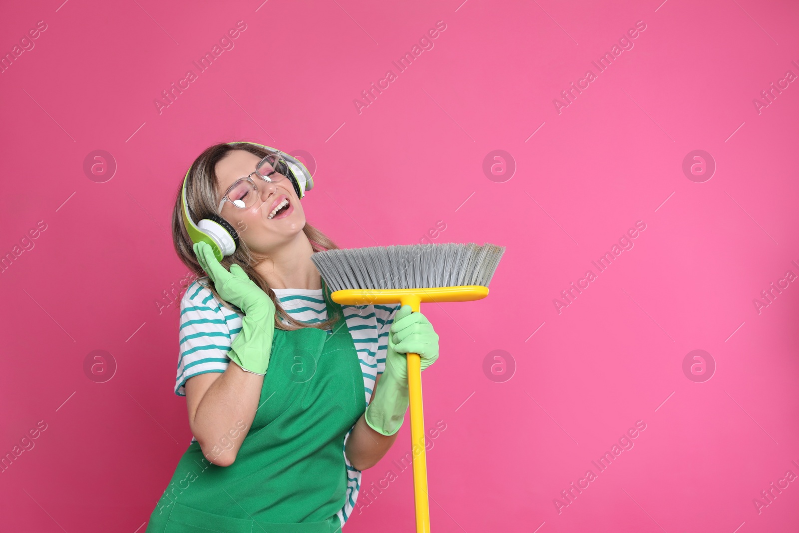 Photo of Beautiful young woman with headphones and floor brush singing on pink background. Space for text