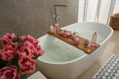 Photo of Wooden bath tray with candles and personal care products on tub indoors