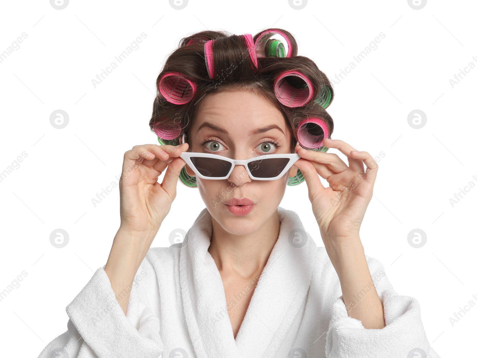 Photo of Emotional young woman in bathrobe with hair curlers and sunglasses on white background