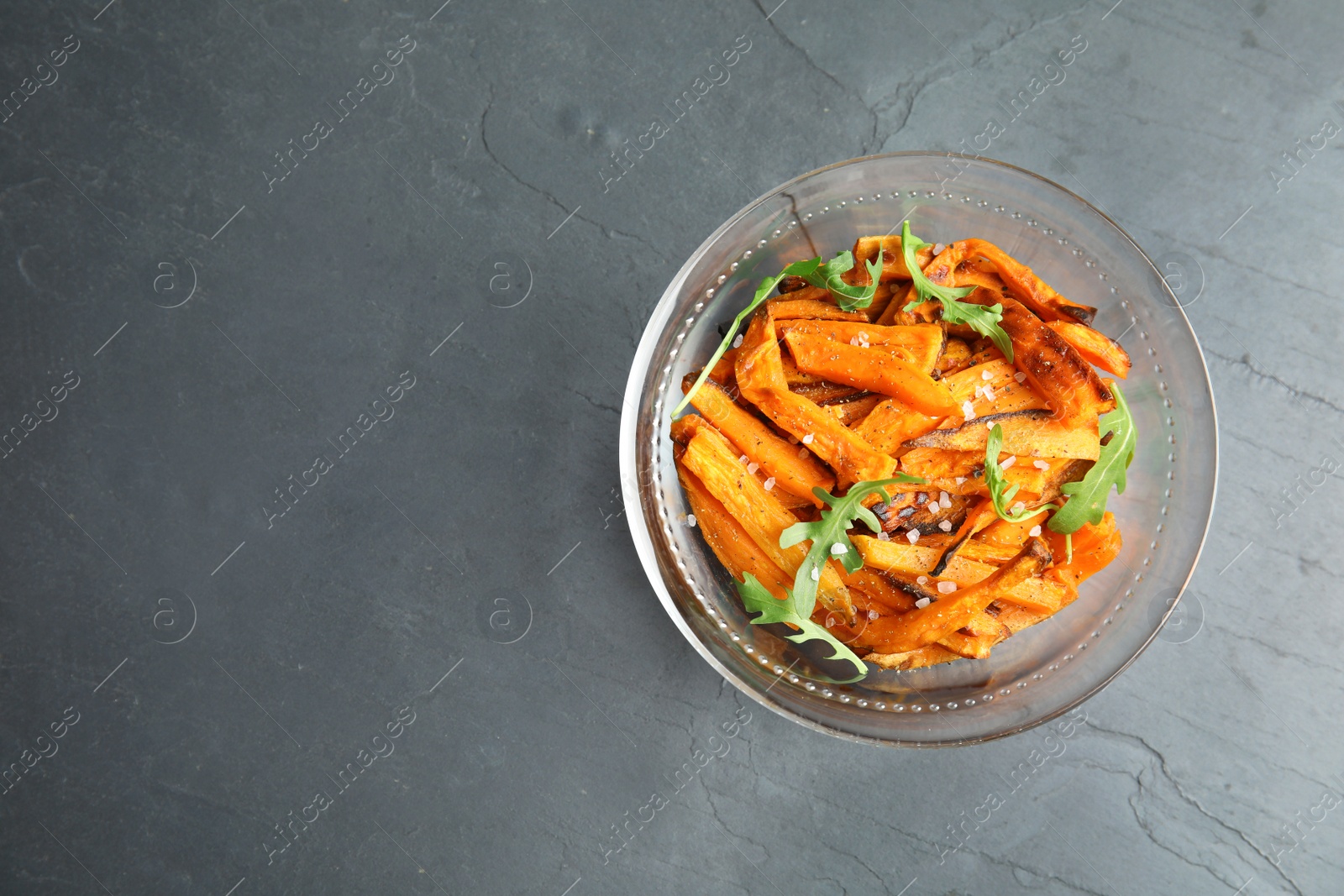 Photo of Glass bowl with baked sweet potato slices and arugula on grey background, top view. Space for text