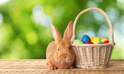 Adorable bunny and wicker basket with Easter eggs on wooden surface outdoors, space for text