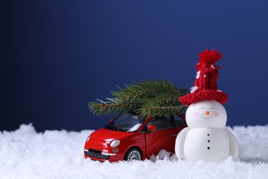 Photo of Cute decorative snowman and toy car with fir tree branches on artificial snow against blue background, space for text