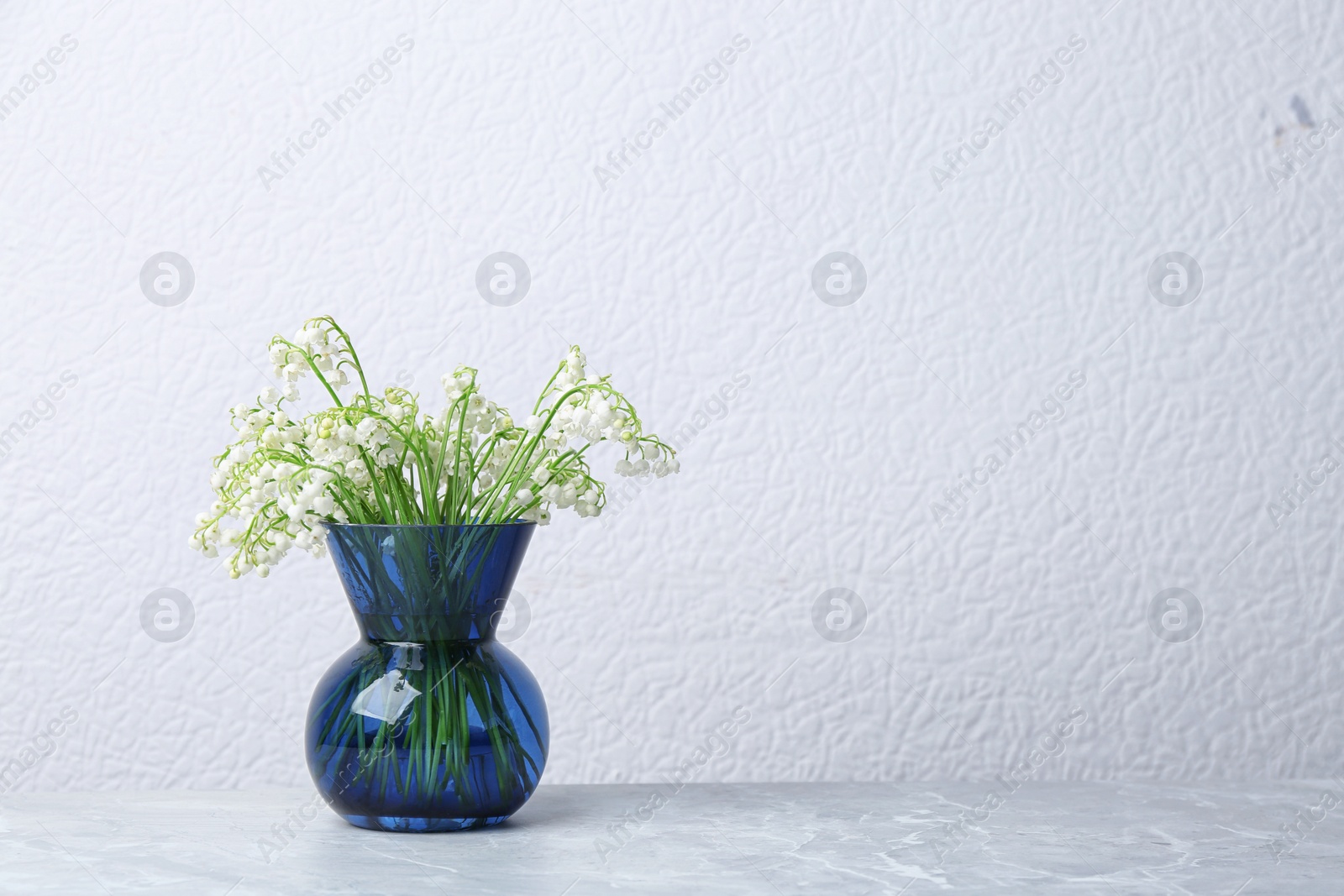 Photo of Beautiful lily of the valley bouquet in vase on table near white wall, space for text