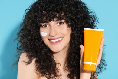 Photo of Beautiful young woman with sun protection cream on her face holding tube of sunscreen against light blue background