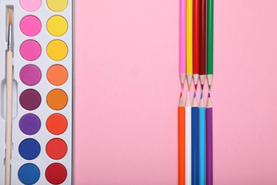 Photo of Watercolor palette with brush and colorful pencils on pink background, flat lay. Space for text