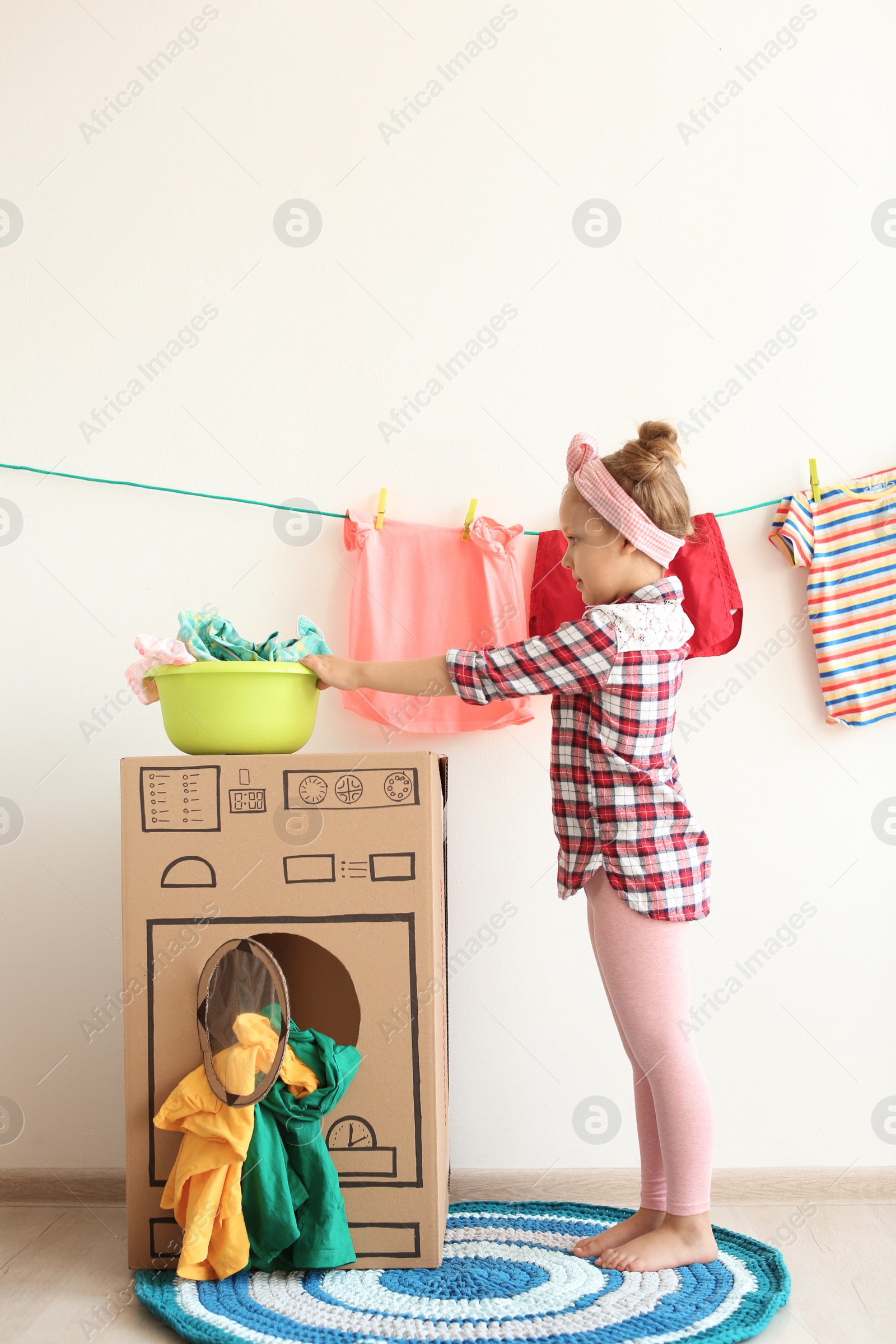 Photo of Adorable little child playing with cardboard washing machine and clothes indoors