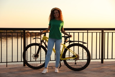 Photo of Beautiful young African-American woman with bicycle on city waterfront at sunset