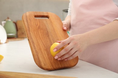Photo of Woman rubbing wooden cutting board with lemon at white table in kitchen, closeup