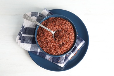 Photo of Bowl with uncooked red rice and spoon on white table, top view