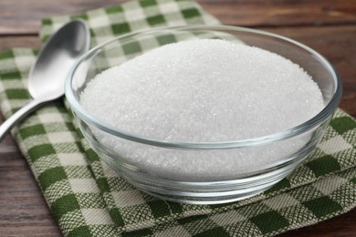 Photo of Granulated sugar in bowl and napkin on wooden table, closeup