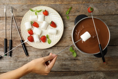 Woman dipping marshmallow into pot with chocolate fondue on wooden background, flat lay