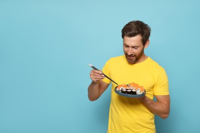Photo of Happy man holding plate with tasty sushi rolls and chopsticks on light blue background. Space for text