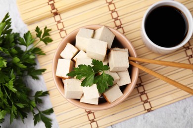 Photo of Bamboo mat with bowl of smoked tofu cubes, soy sauce and parsley on white textured table, flat lay