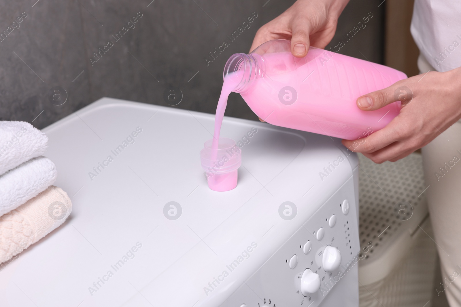 Photo of Woman pouring fabric softener from bottle into cap on washing machine indoors, closeup