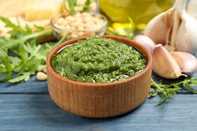 Photo of Bowl of tasty arugula pesto and ingredients on blue wooden table