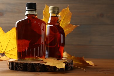 Photo of Bottles of tasty maple syrup and dry leaves on wooden table, space for text