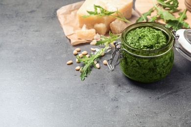 Photo of Jar of tasty arugula pesto and ingredients on grey table. Space for text