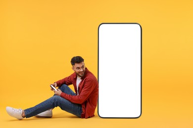 Image of Man with mobile phone sitting near huge device with empty screen on orange background. Mockup for design