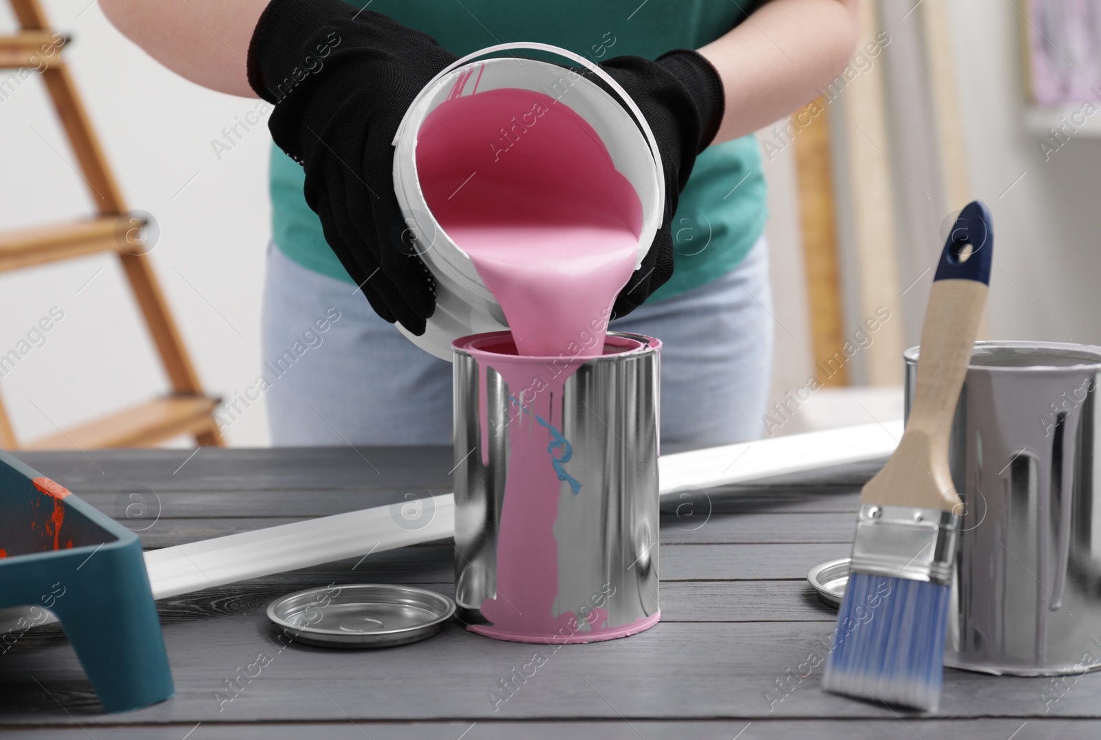 Photo of Woman pouring pink paint from bucket into can at grey wooden table indoors, closeup