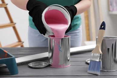 Woman pouring pink paint from bucket into can at grey wooden table indoors, closeup