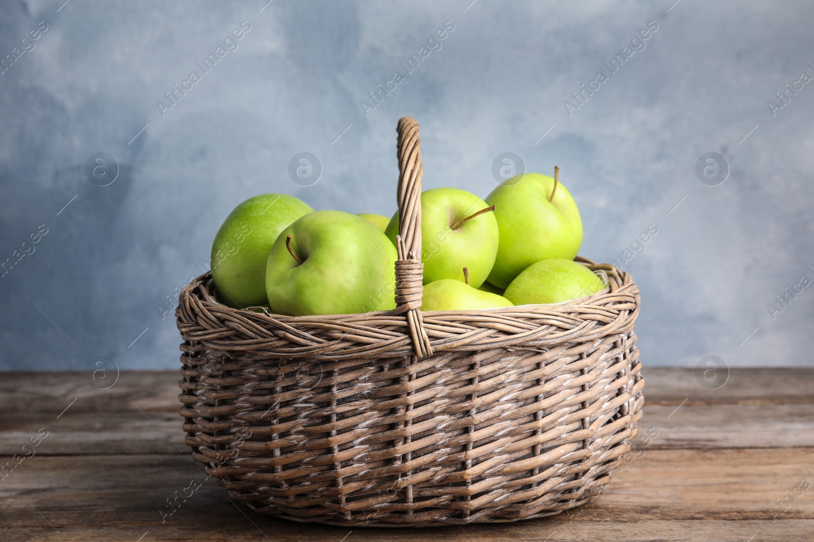 Photo of Wicker basket of fresh ripe green apples on wooden table against blue background