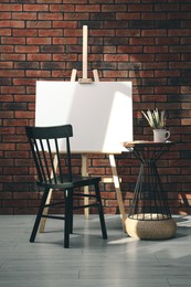 Wooden easel with blank canvas and different art supplies near brick wall in room