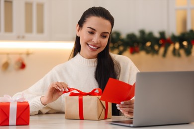 Photo of Celebrating Christmas online with exchanged by mail presents. Smiling woman reading greeting card and opening gift during video call at home