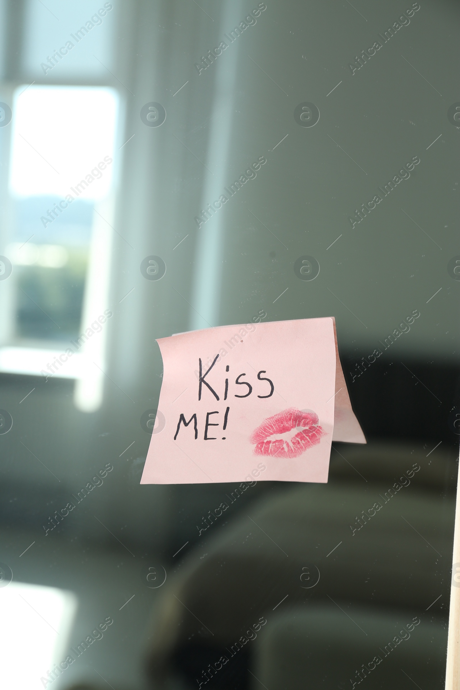Photo of Sticky note with phrase Kiss Me and lipstick mark attached to mirror in room