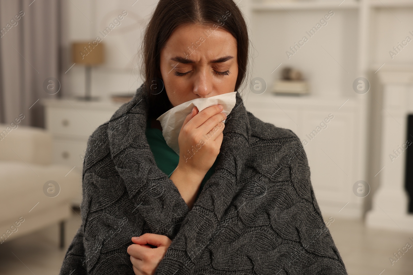 Photo of Sick woman wrapped in blanket with tissue blowing nose at home. Cold symptoms