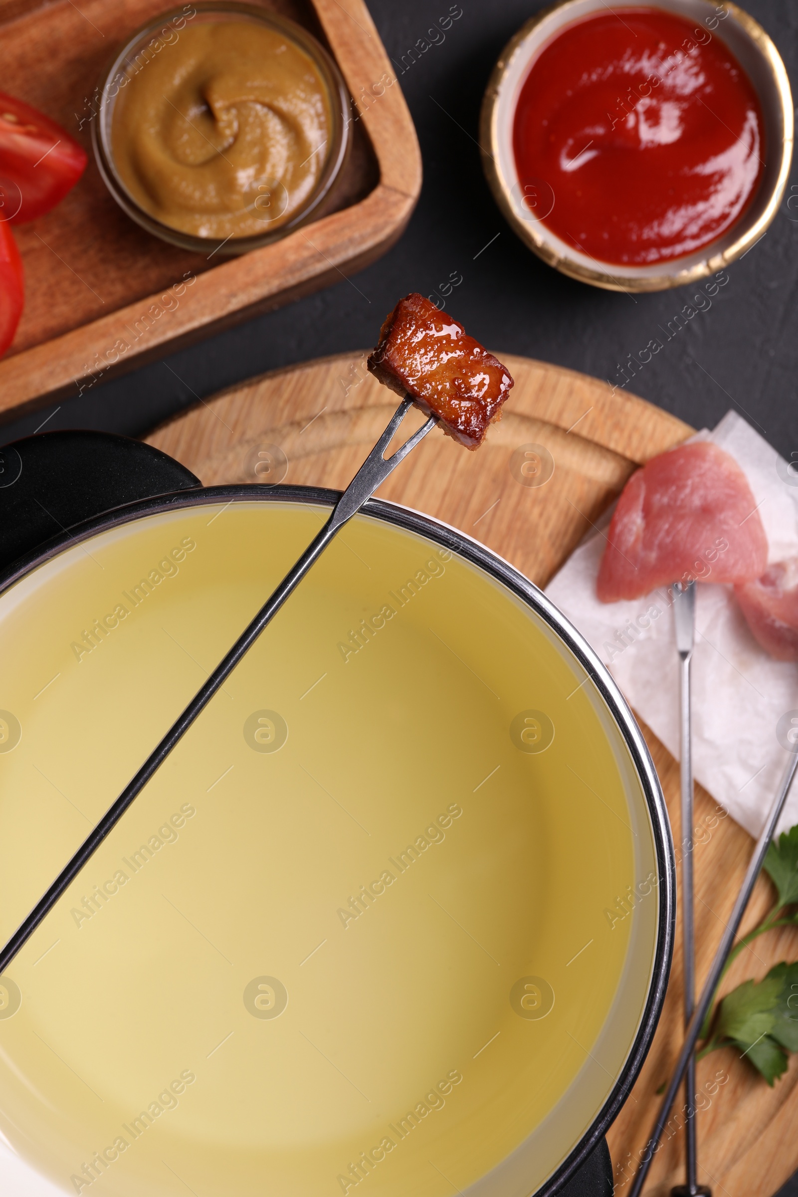 Photo of Fondue pot, forks with meat pieces and other products on black textured table, flat lay