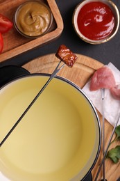 Photo of Fondue pot, forks with meat pieces and other products on black textured table, flat lay