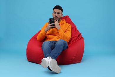 Handsome young man using smartphone on bean bag chair against light blue background