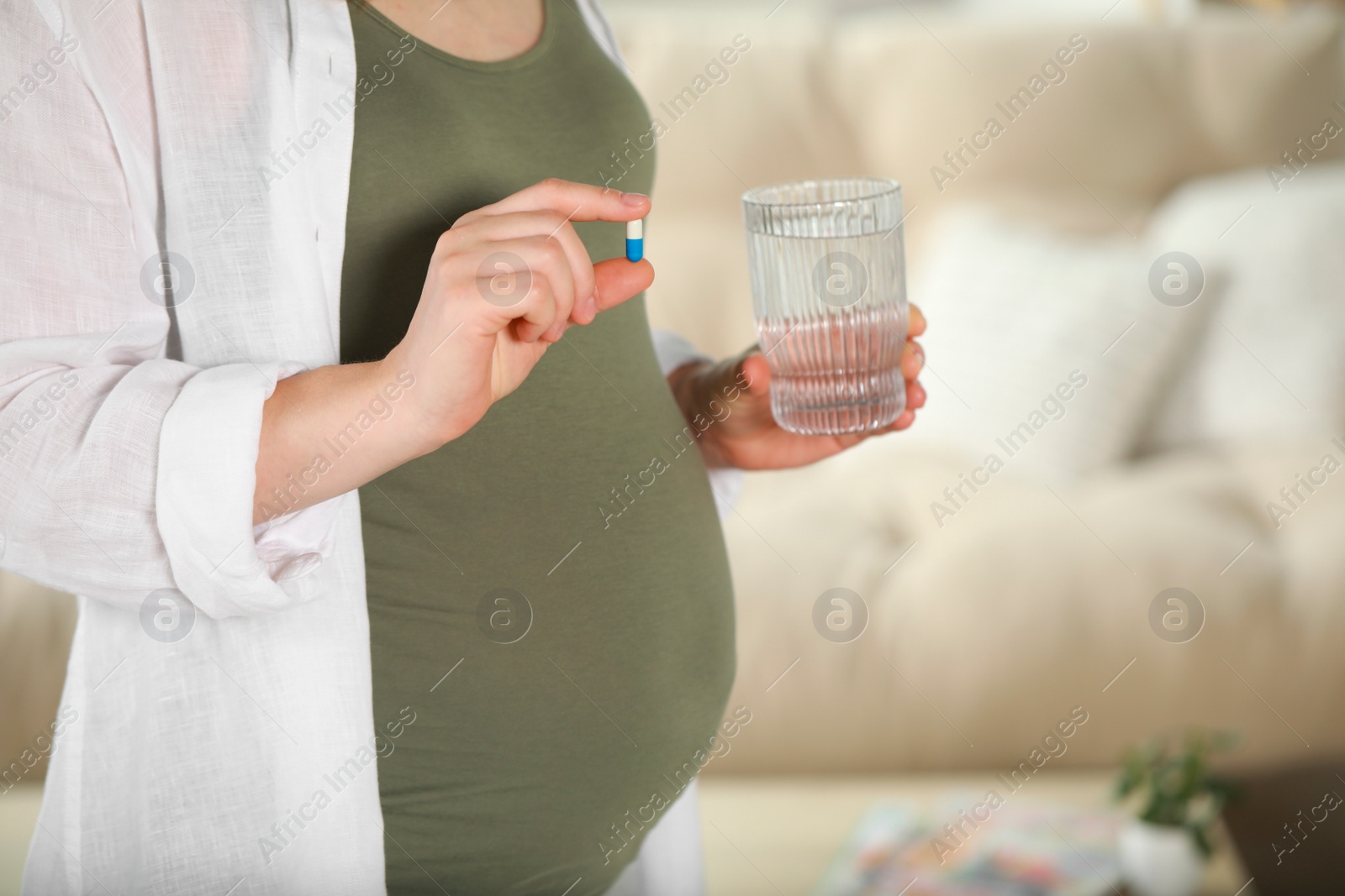 Photo of Pregnant woman holding pill and glass of water near sofa indoors, closeup