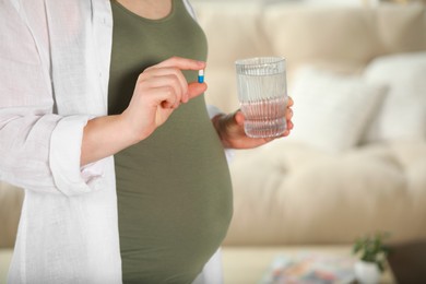 Pregnant woman holding pill and glass of water near sofa indoors, closeup
