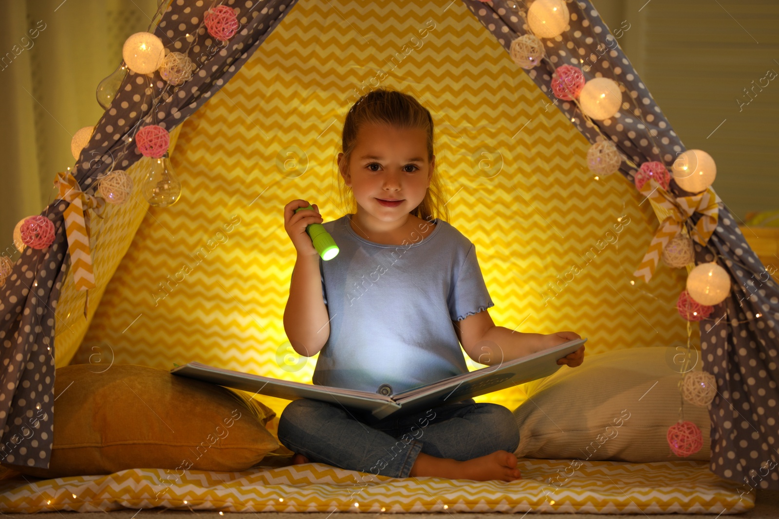 Photo of Little girl with flashlight reading book in play tent at home