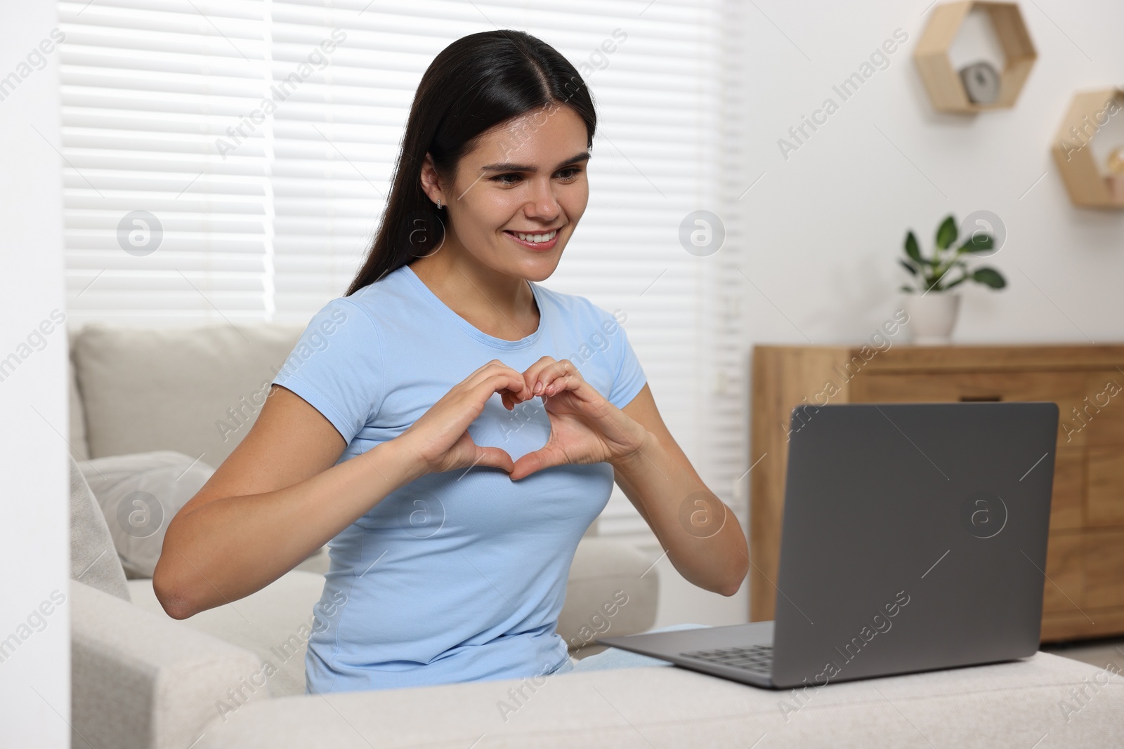 Photo of Happy young woman having video chat via laptop and making heart with hands on sofa in living room