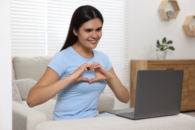Happy young woman having video chat via laptop and making heart with hands on sofa in living room