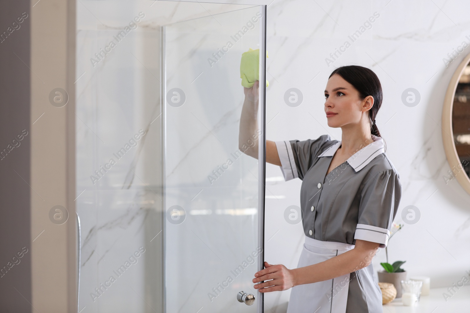 Photo of Young chambermaid wiping dust from shower booth in bathroom