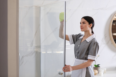 Young chambermaid wiping dust from shower booth in bathroom
