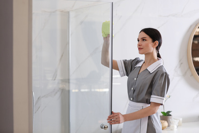 Photo of Young chambermaid wiping dust from shower booth in bathroom