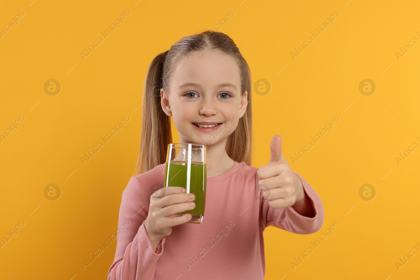 Photo of Cute little girl with glass of fresh juice showing thumbs up on orange background