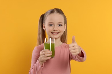 Cute little girl with glass of fresh juice showing thumbs up on orange background