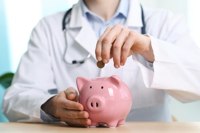 Photo of Doctor putting coin into piggy bank at wooden table, closeup