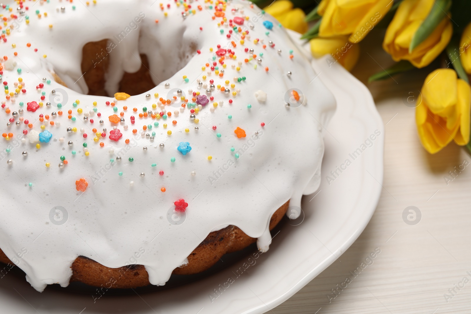 Photo of Easter cake with sprinkles and tulips on white wooden table, closeup