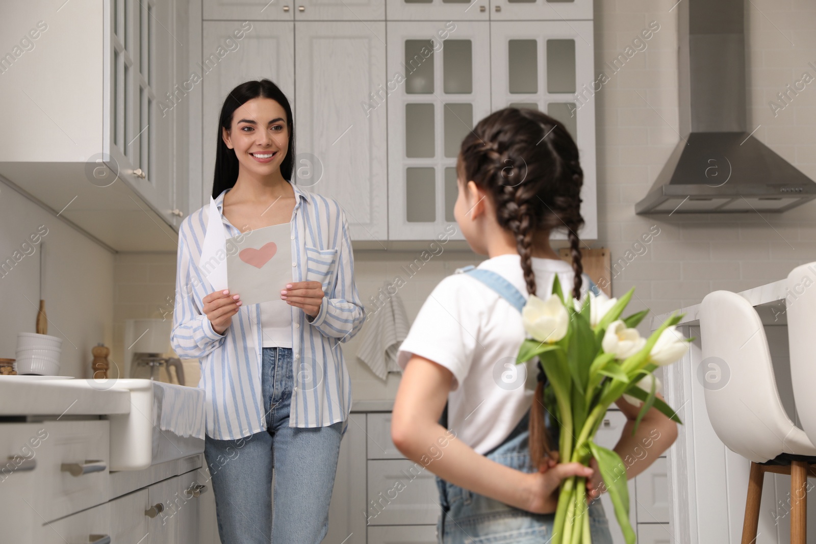 Photo of Little girl hiding tulip bouquet for mom in kitchen at home. Happy Mother's Day