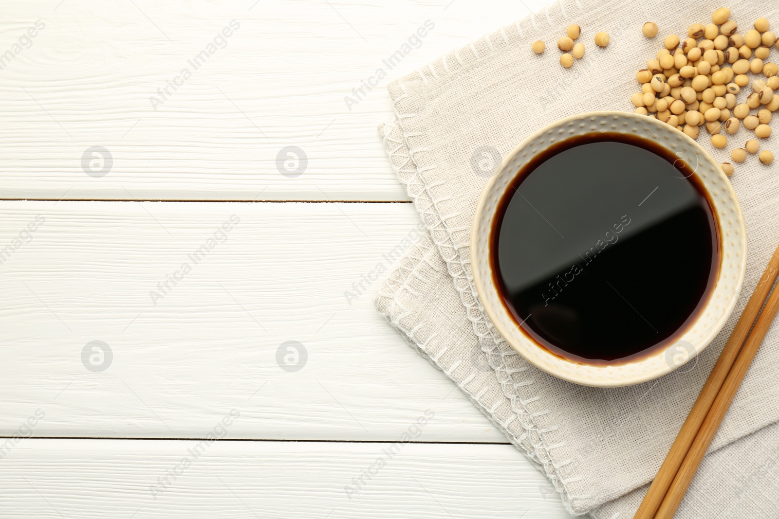 Photo of Tasty soy sauce in bowl, chopsticks and soybeans on white wooden table, top view. Space for text