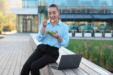 Smiling businesswoman eating from lunch box near laptop outdoors
